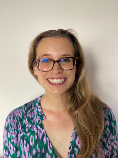 Smiling person with glasses in a patterned green-and-pink blouse against a plain wall