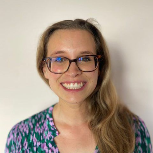 Smiling person with glasses and long hair in a patterned top against a plain background