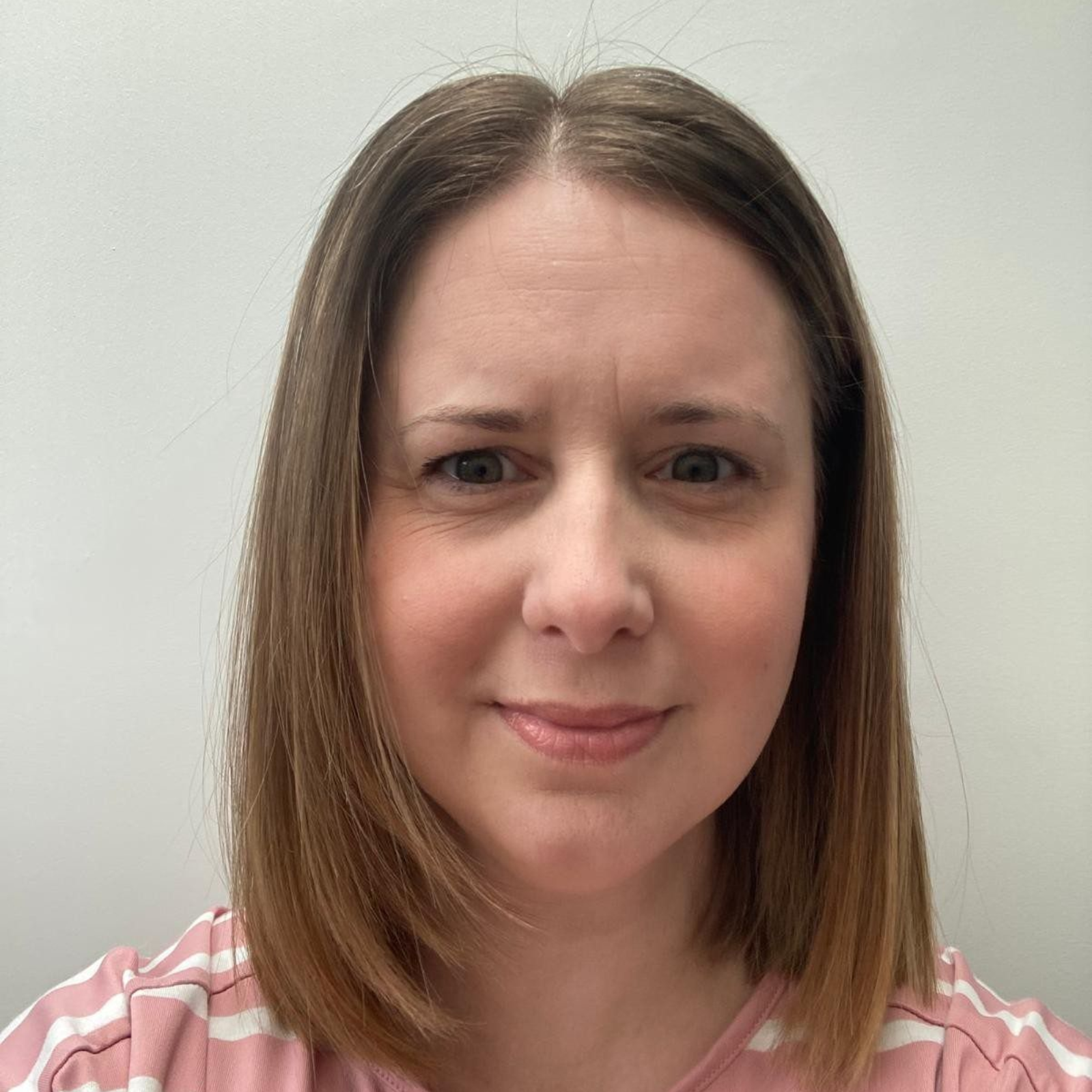 Close-up portrait of a woman with shoulder-length brown hair, wearing a pink striped shirt, against a plain wall