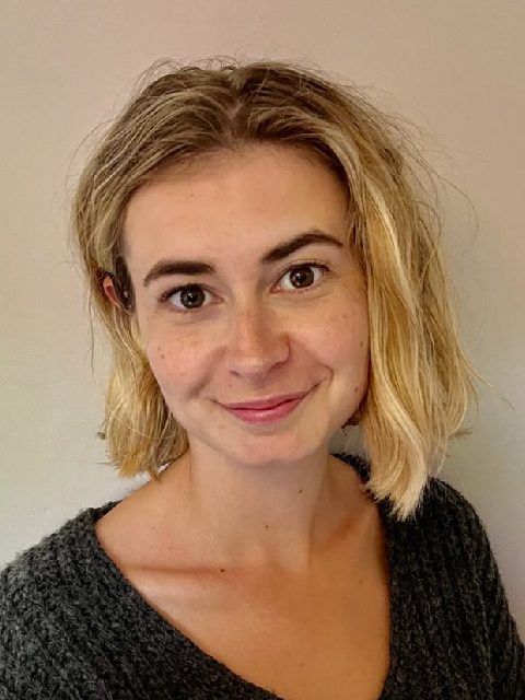 Smiling woman with blonde hair in a dark sweater against a plain beige wall