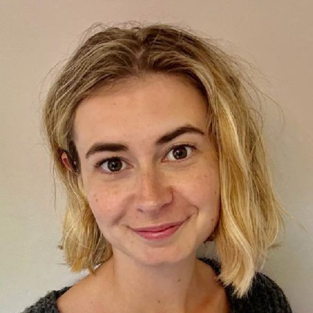 Smiling person with short blonde hair against a plain beige background