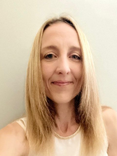 Close-up portrait of a smiling person with straight blonde hair against a plain light background