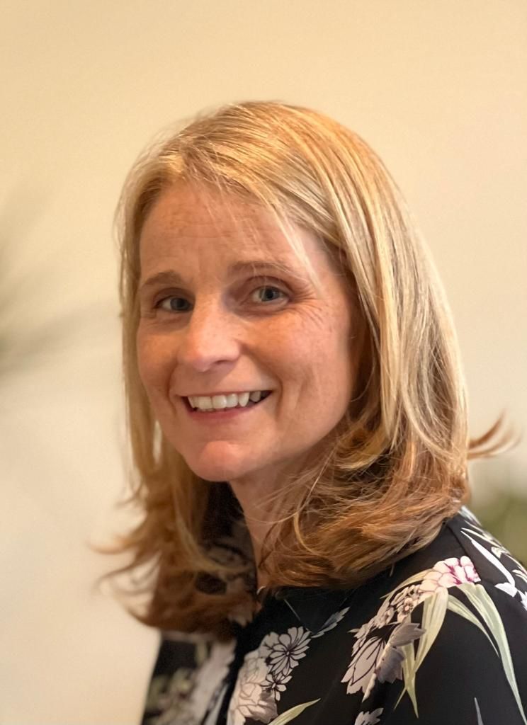 Smiling blonde woman in a black floral blouse against a beige wall
