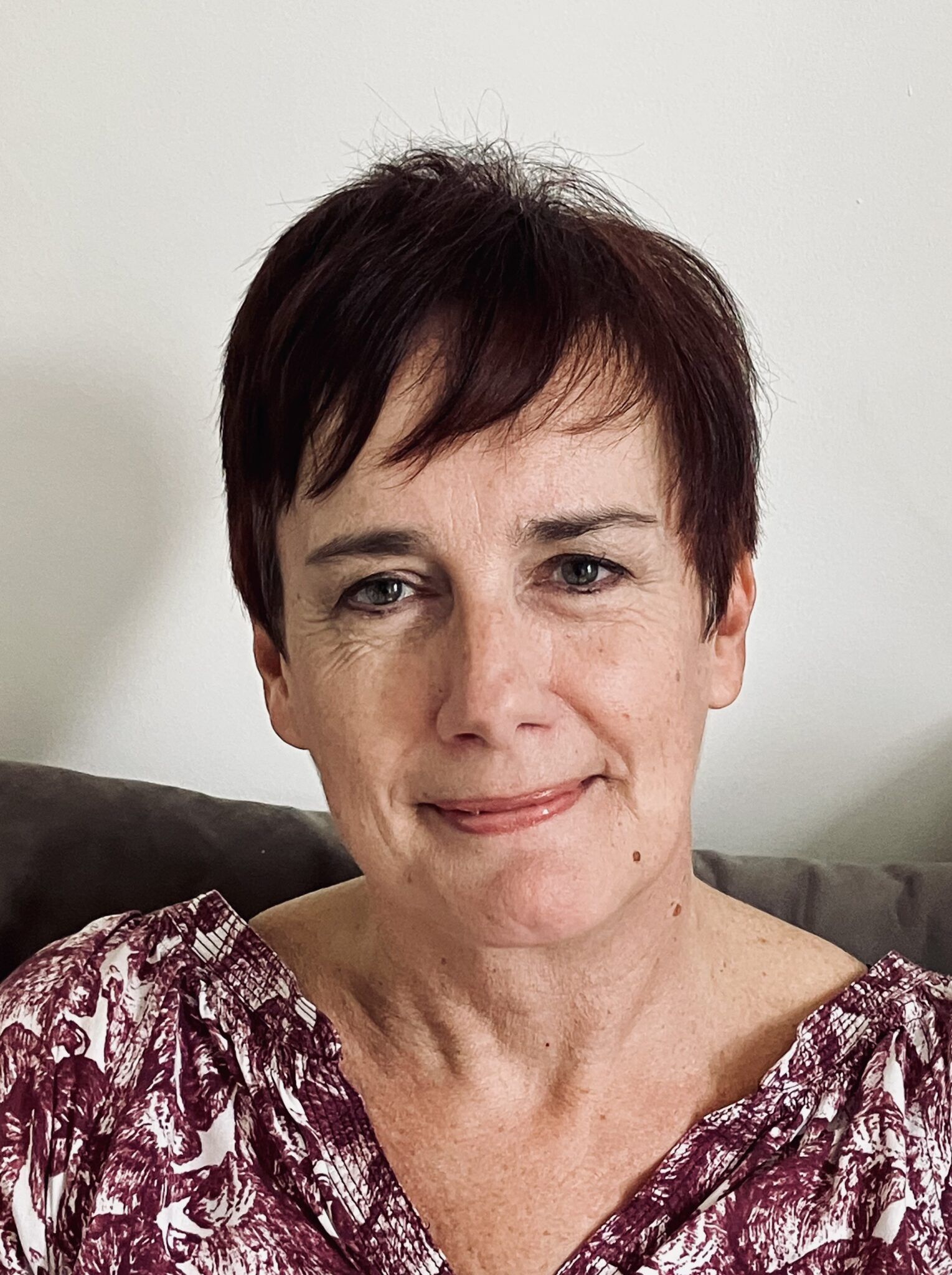 Portrait of a smiling woman with short dark hair, wearing a patterned top, seated indoors on a couch