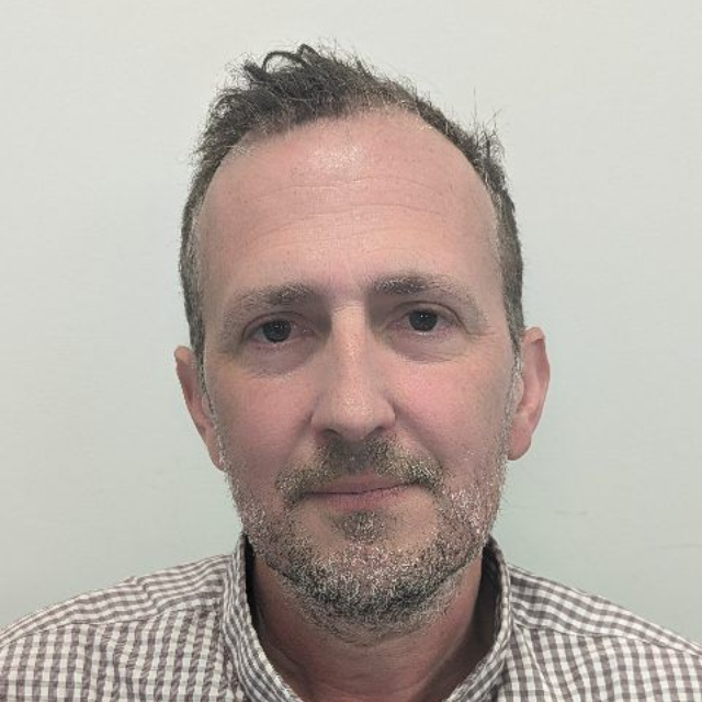 Portrait of a man with short dark hair and stubble, wearing a plaid shirt against a plain light background