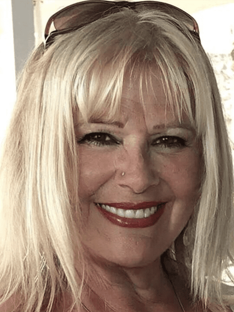 Smiling blonde woman with bangs and sunglasses on her head, wearing red lipstick.