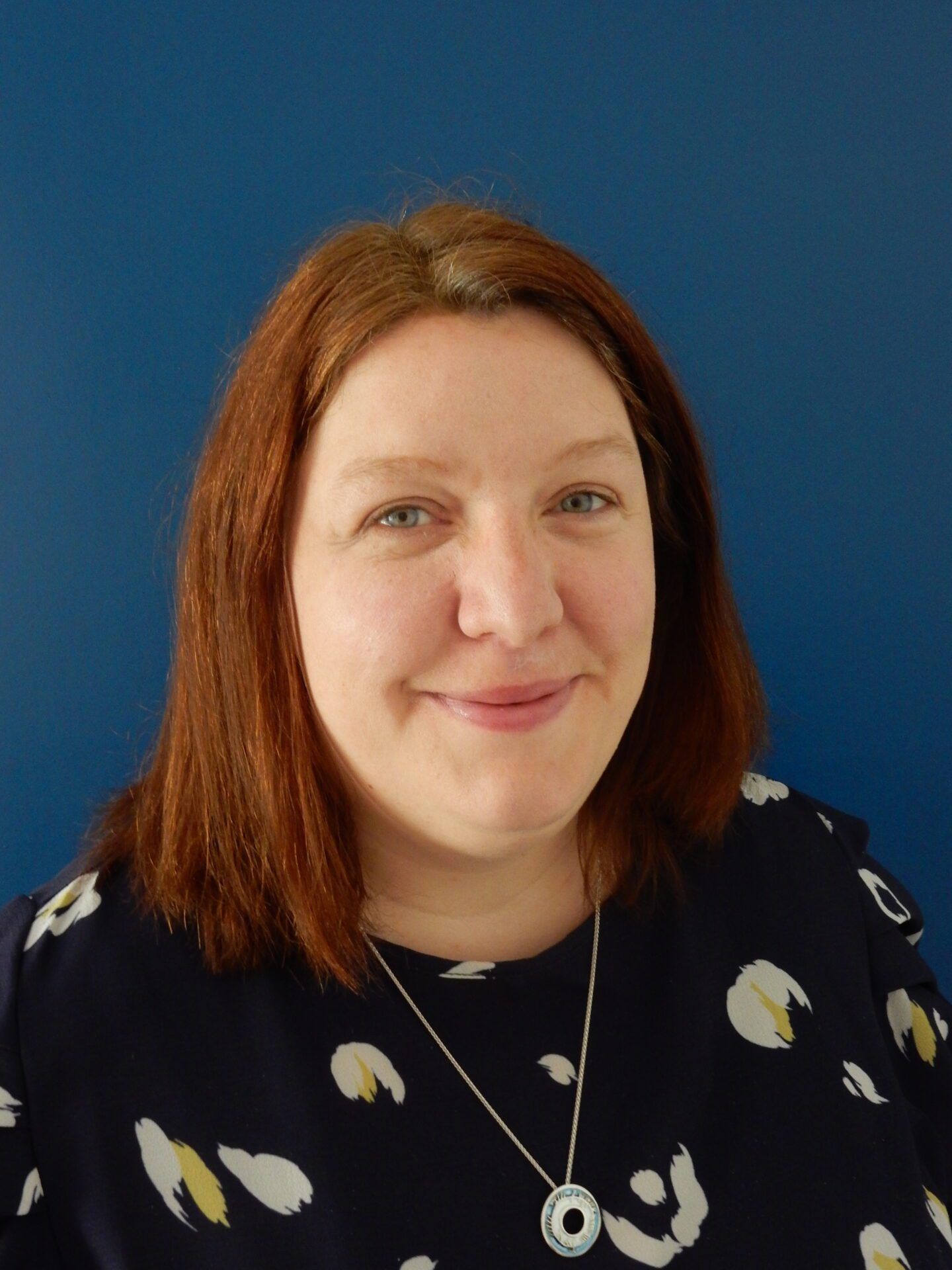 Portrait of a smiling person with red hair, wearing a dark patterned top against a blue background
