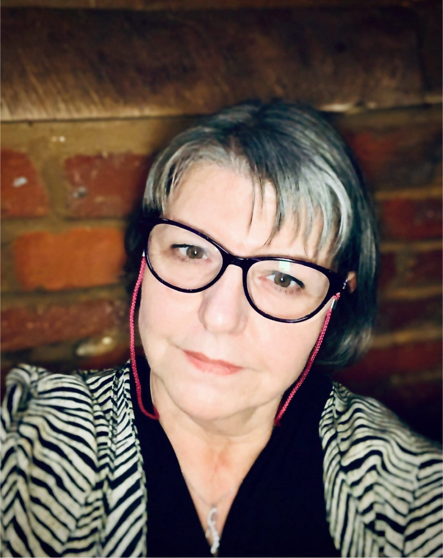Portrait of a person wearing black glasses and a zebra-pattern top, seated against a brick wall