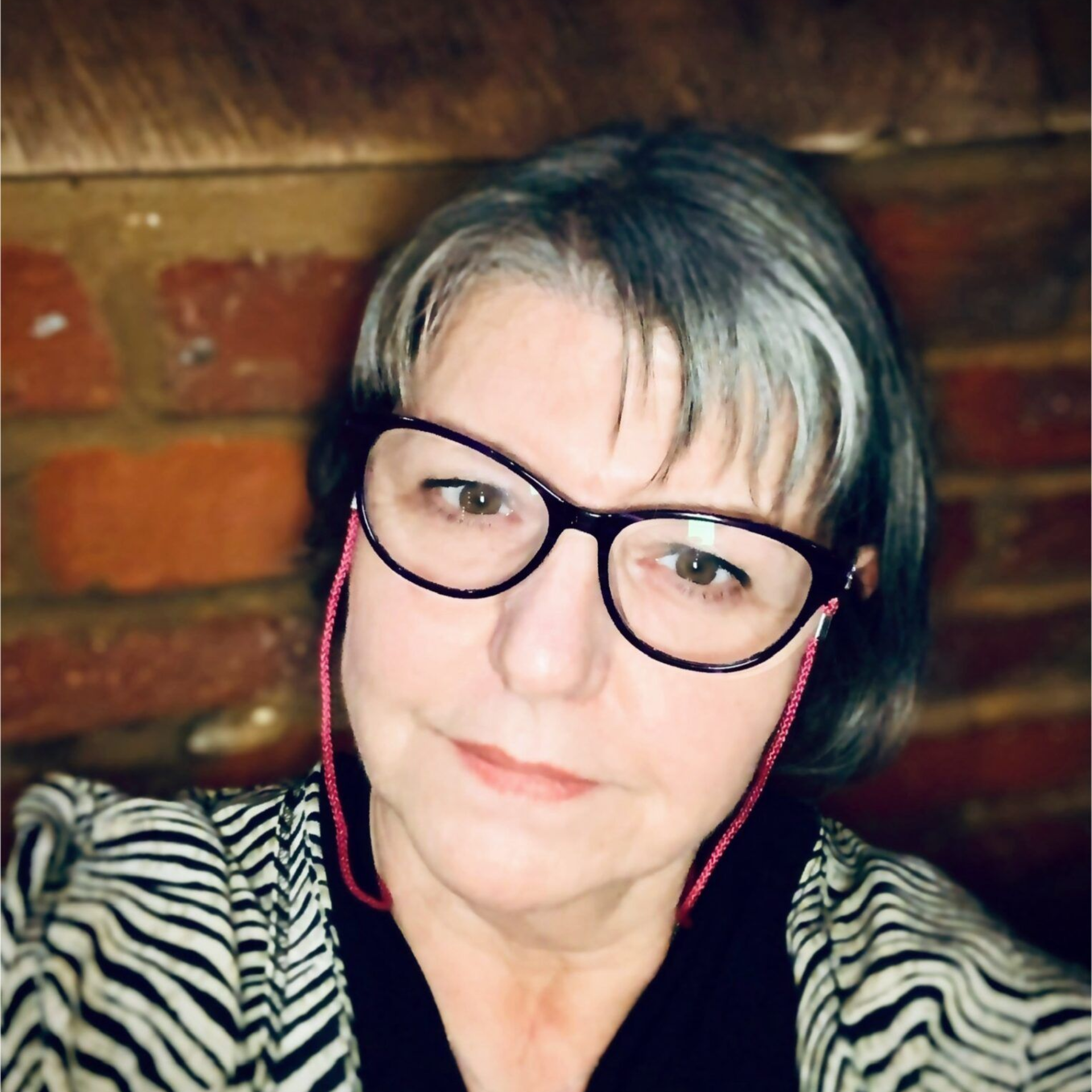 Close-up selfie of a person wearing black glasses and a zebra-print top against a wooden wall.