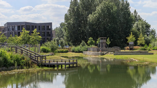 Modern building beside a calm pond with a wooden footbridge and lush green trees under a cloudy sky