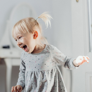 Young child in a gray dress laughing with arms outstretched in a bright room