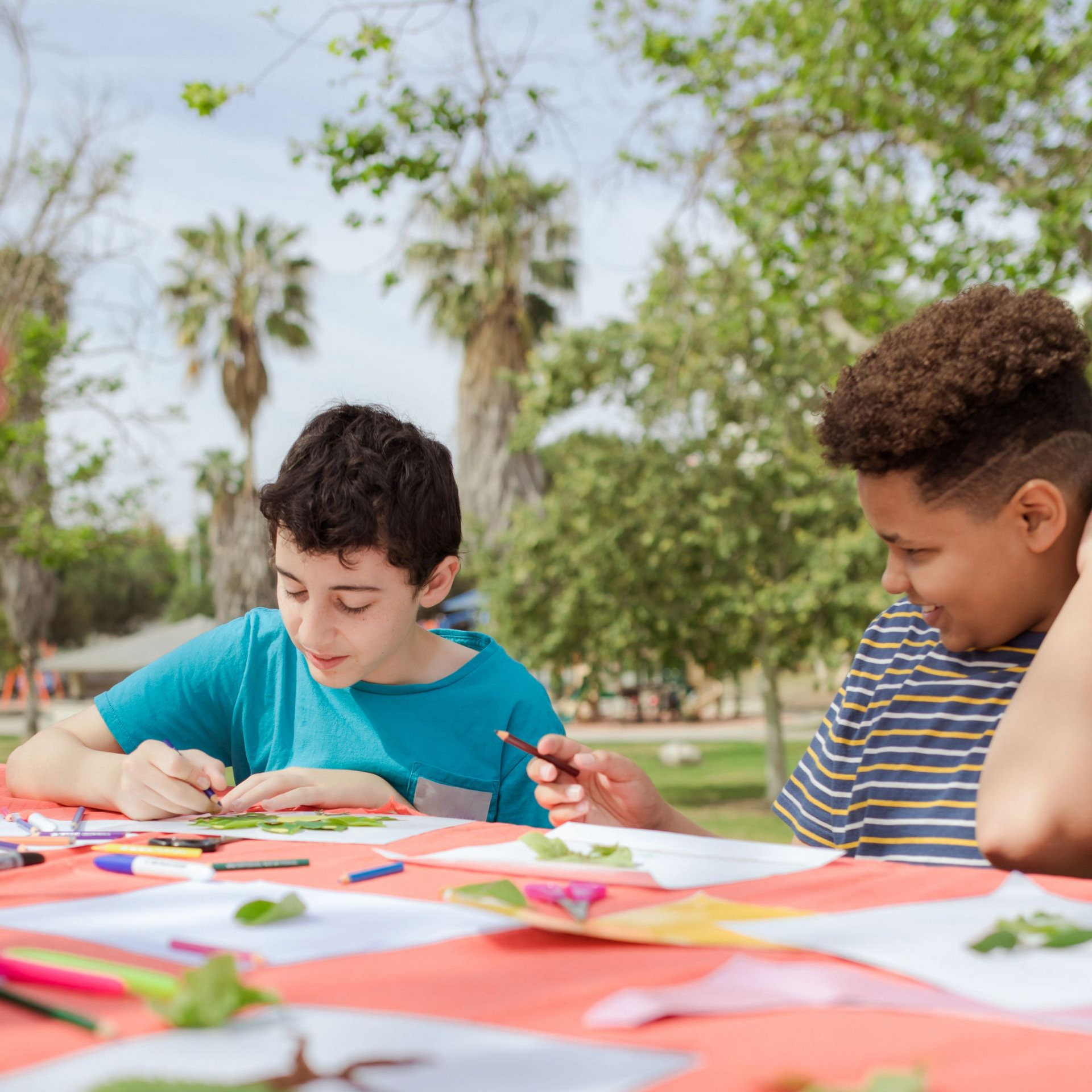 Two children painting at a table outdoors with colorful art supplies and papers