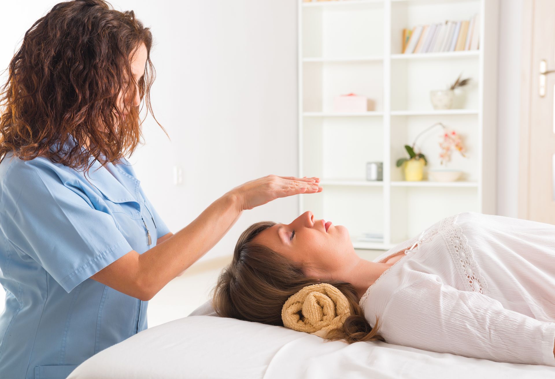 Woman receiving reiki treatment from a practitioner, who hovers her hands over the patient's head in a light-filled room.