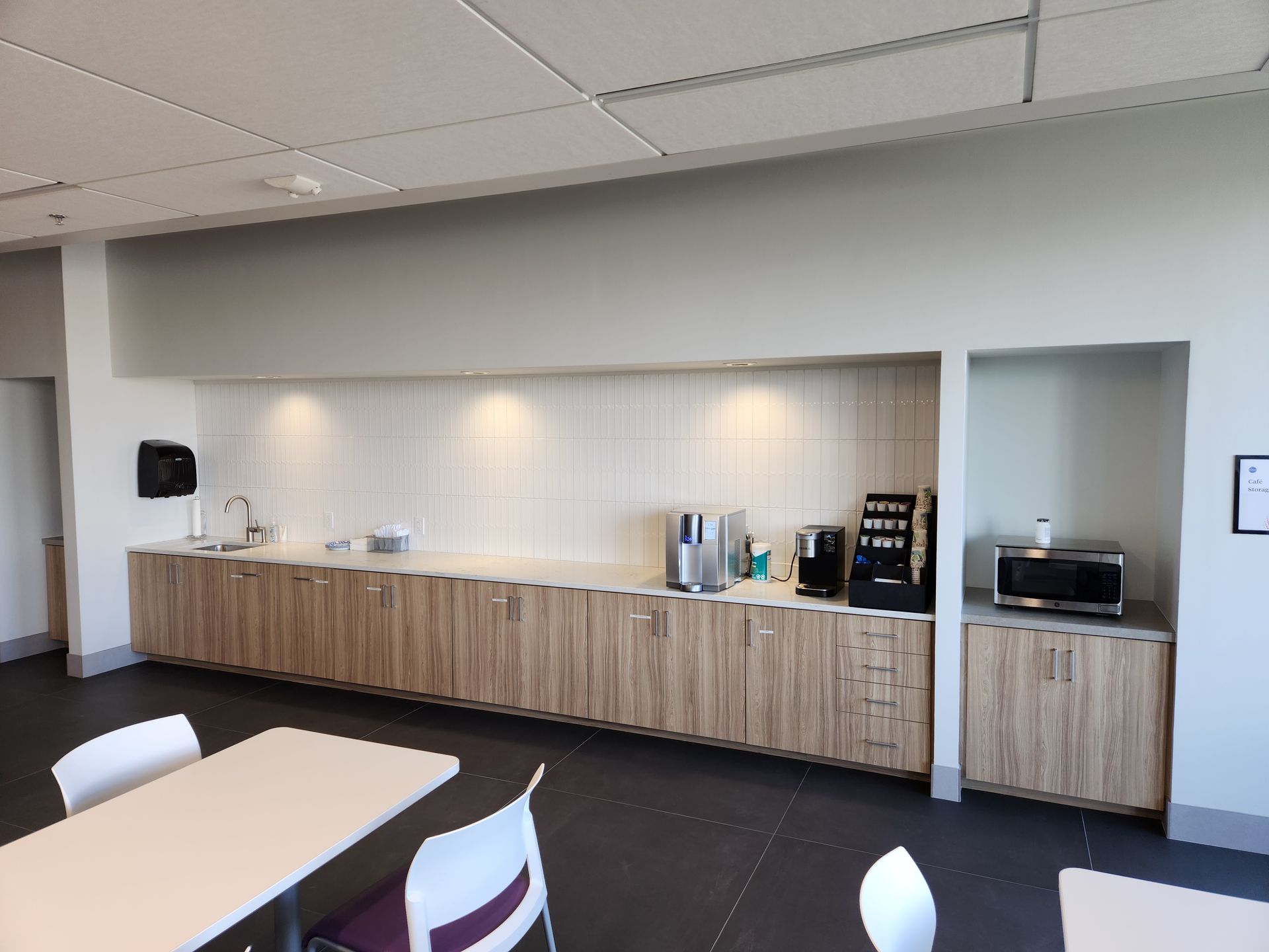 A break room with a long counter, cabinets, coffee maker, and microwave. Tables and chairs are in front.