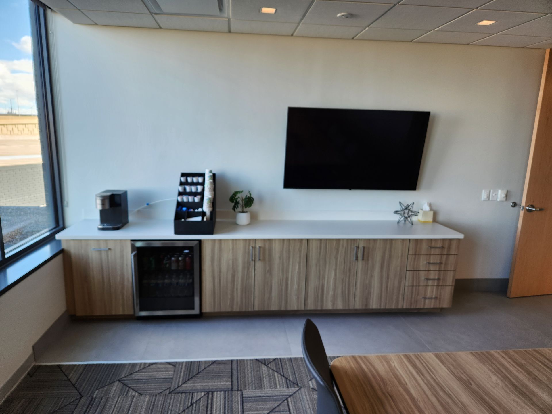 Office breakroom with coffee station, mini fridge, TV, and cabinets against a white wall.