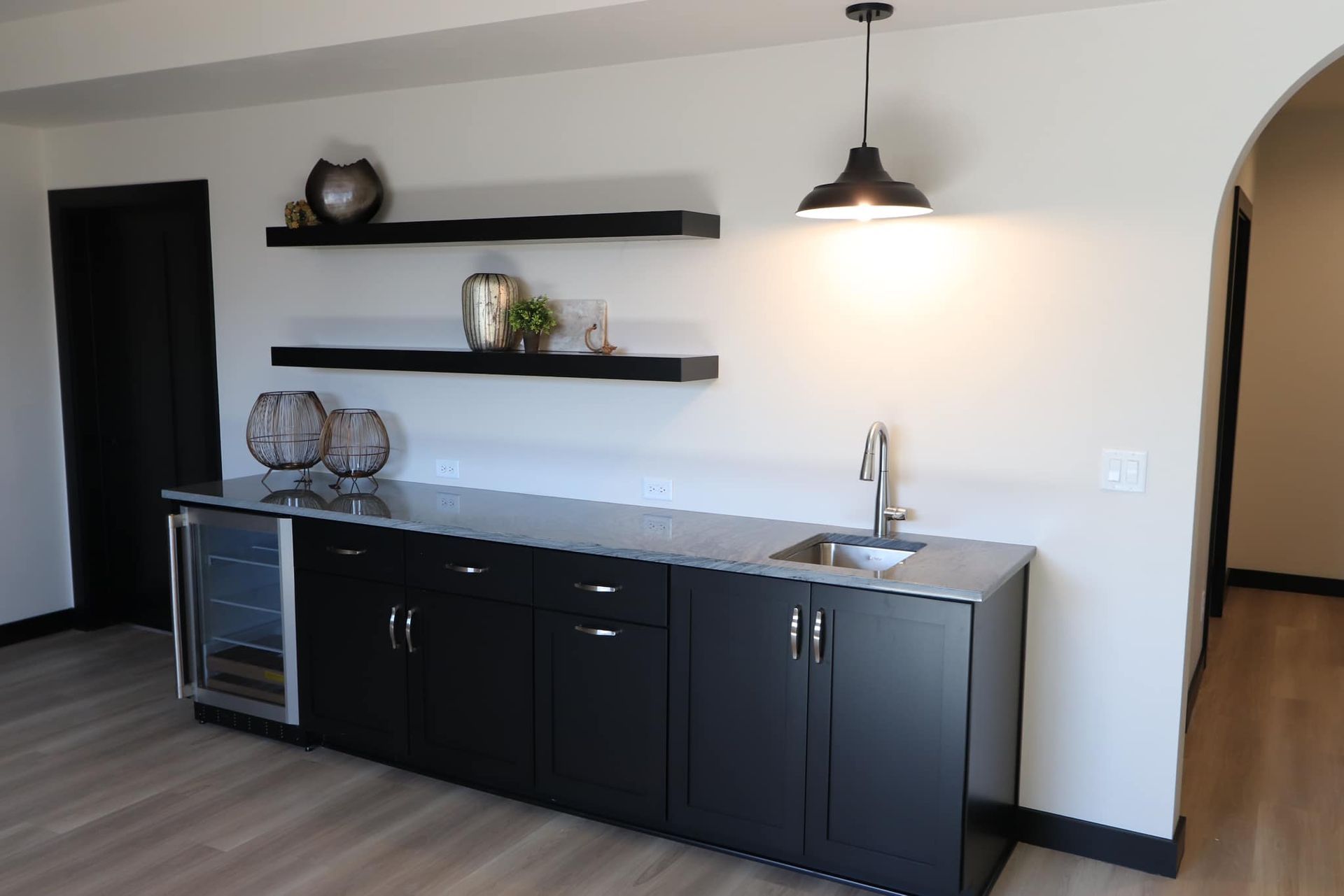 Black home bar with shelves, sink, and wine cooler. Neutral walls, pendant light, arched doorway.
