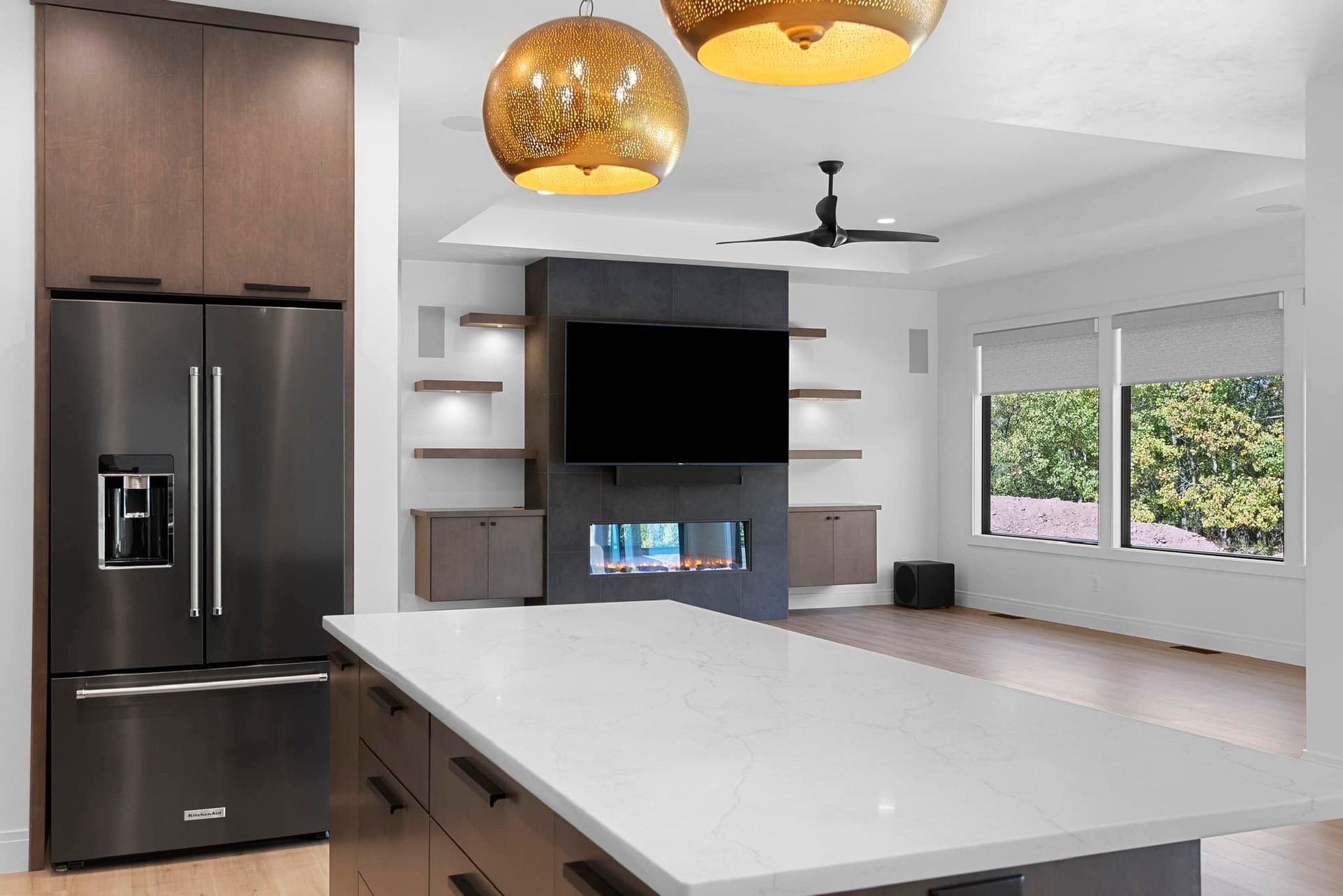Modern kitchen with stainless steel fridge, white island, and dark accent wall with fireplace and TV.