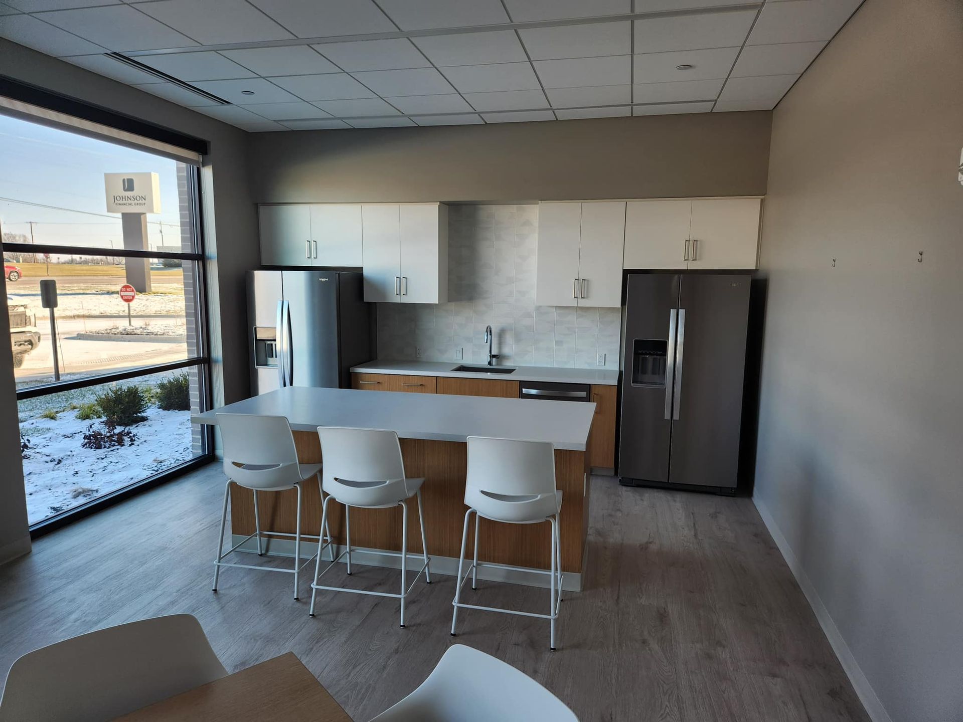 Modern office kitchen with island, white cabinets, two refrigerators, and window overlooking snow.