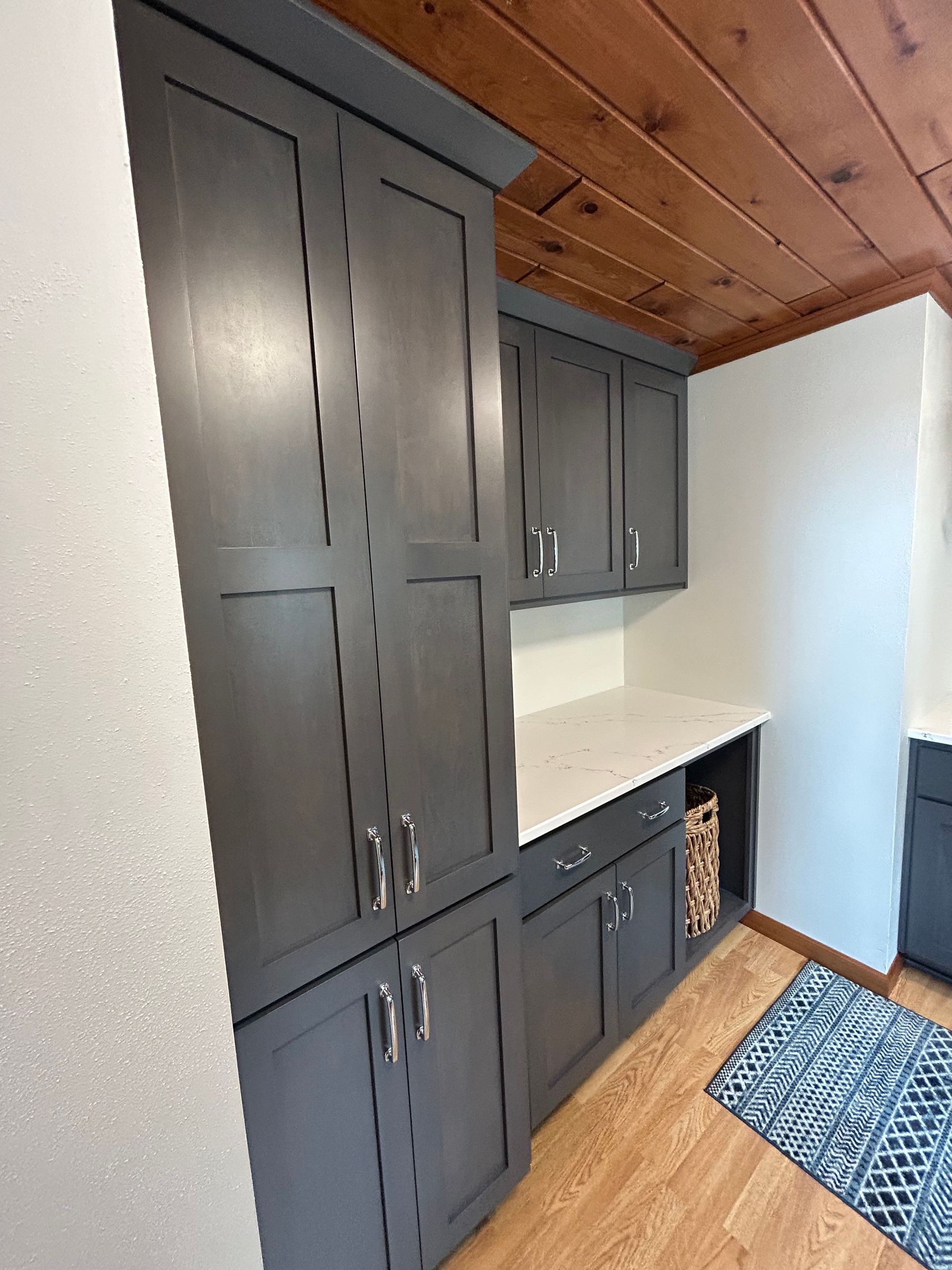 Dark gray cabinets in a kitchen, with a light countertop and wood ceiling, next to white wall and wooden floor.