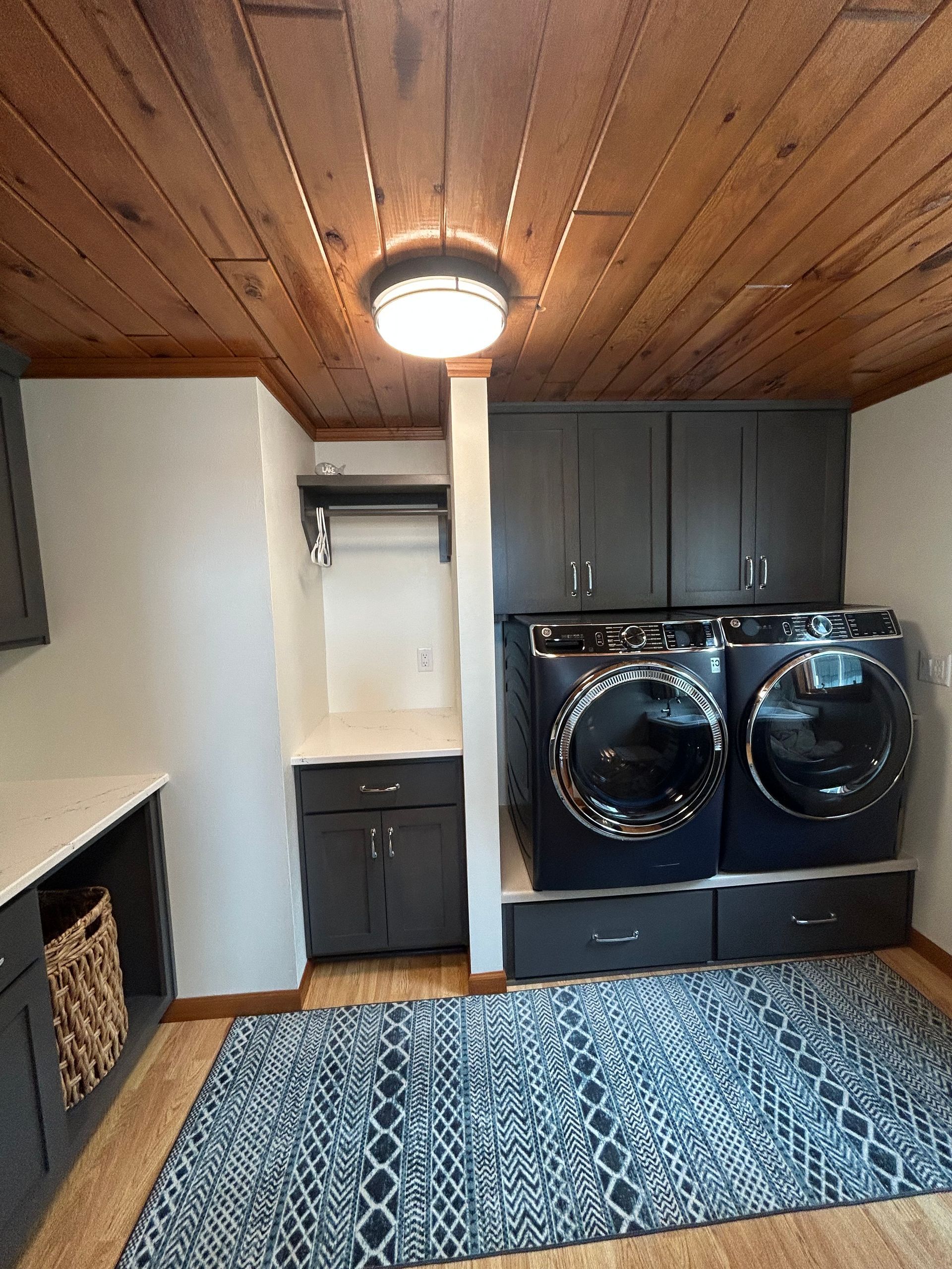 Laundry room with gray cabinets, washer/dryer, wood ceiling, and a patterned rug.
