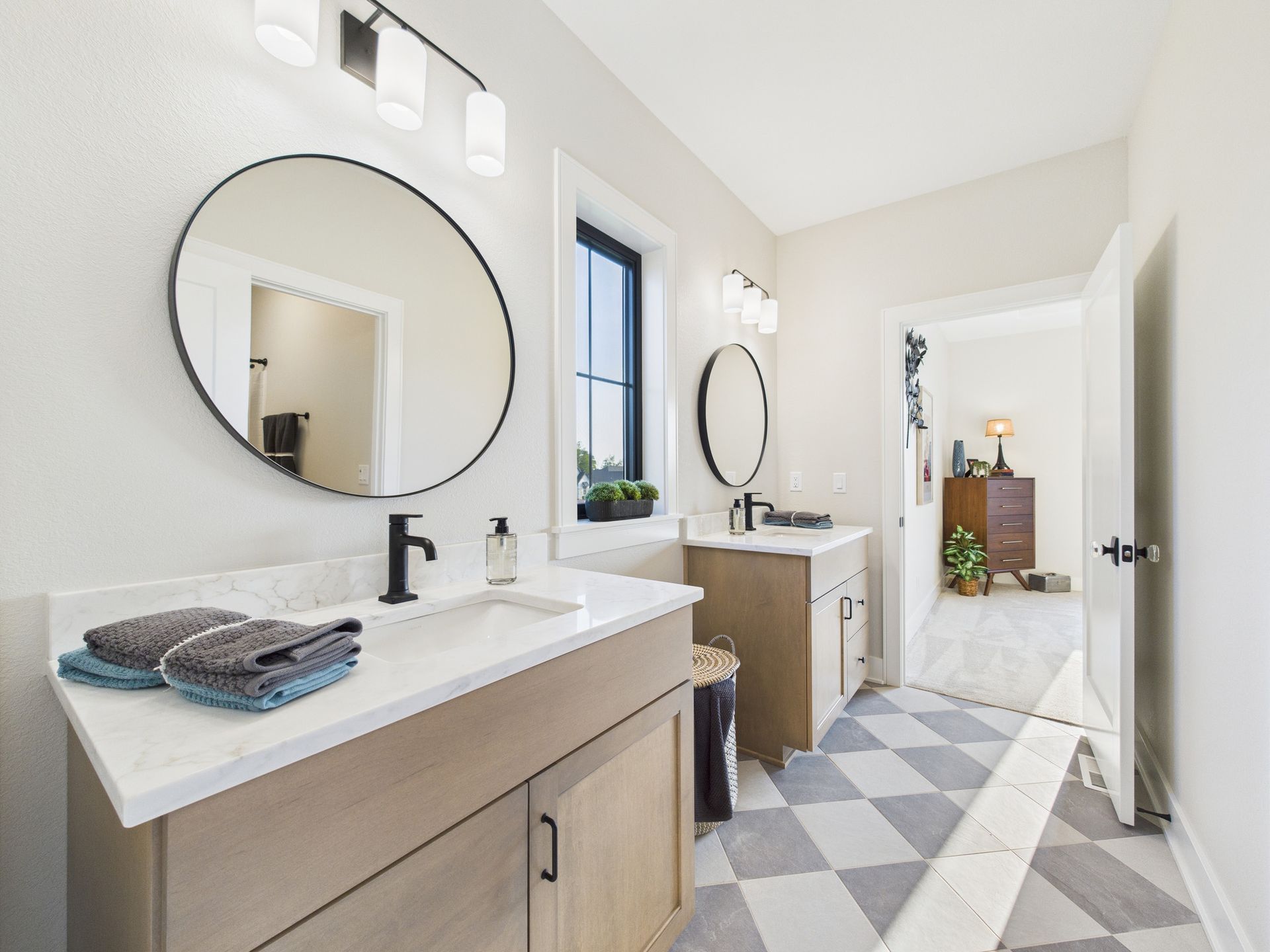 Bathroom with two sinks, round mirrors, and checkered floor. Light wood cabinets, black fixtures, and bright lighting.