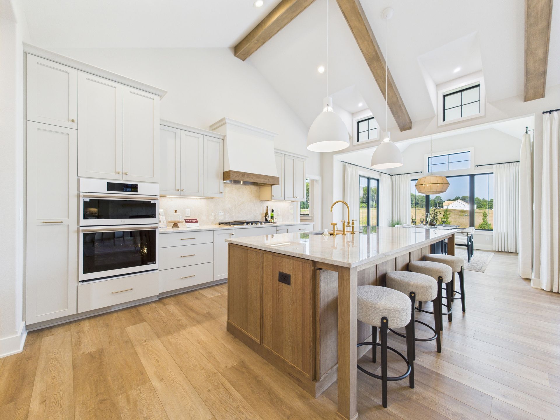 Spacious white kitchen with wooden accents, island, and bar stools. Large windows overlook a green landscape.