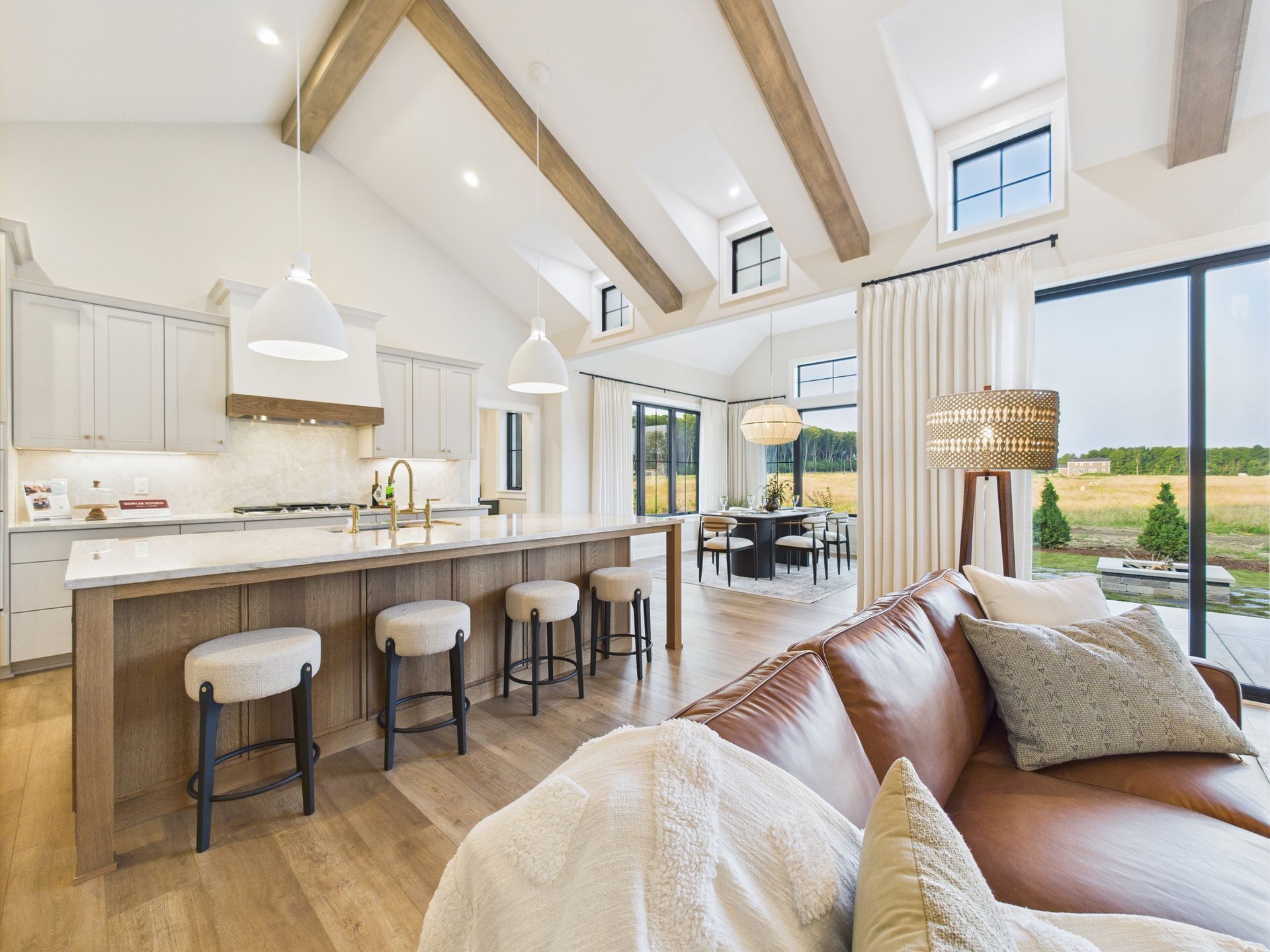 Open-concept kitchen and living space. Light-colored kitchen with island and seating, leather sofa, dining table, and large windows.