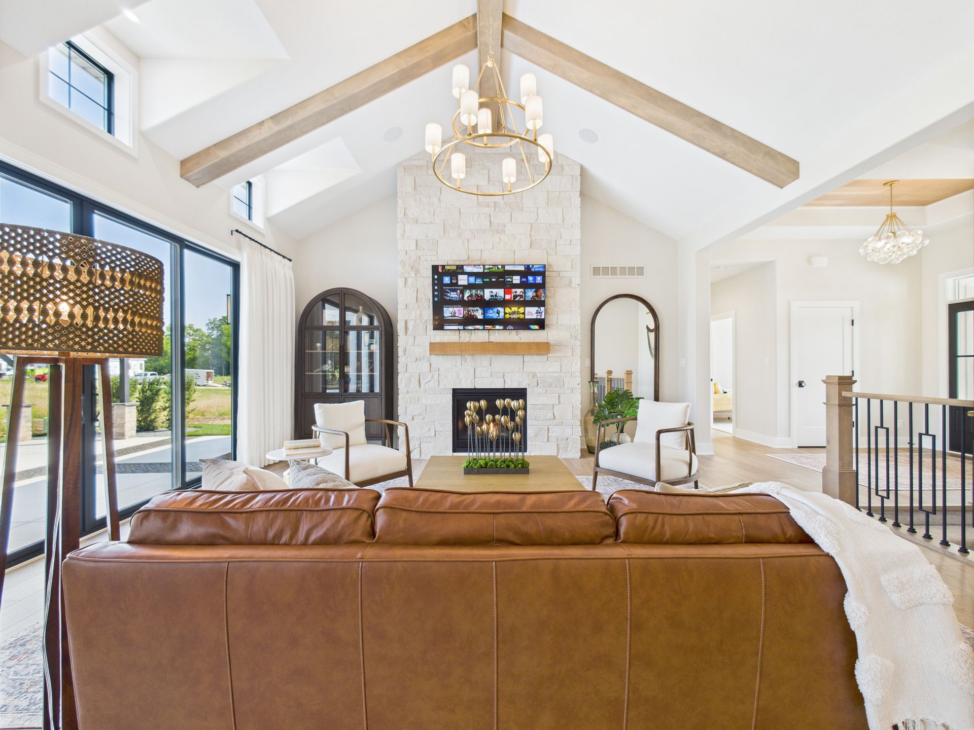 Living room with brown leather sofa, fireplace, large windows, light wood beams, and chandelier.