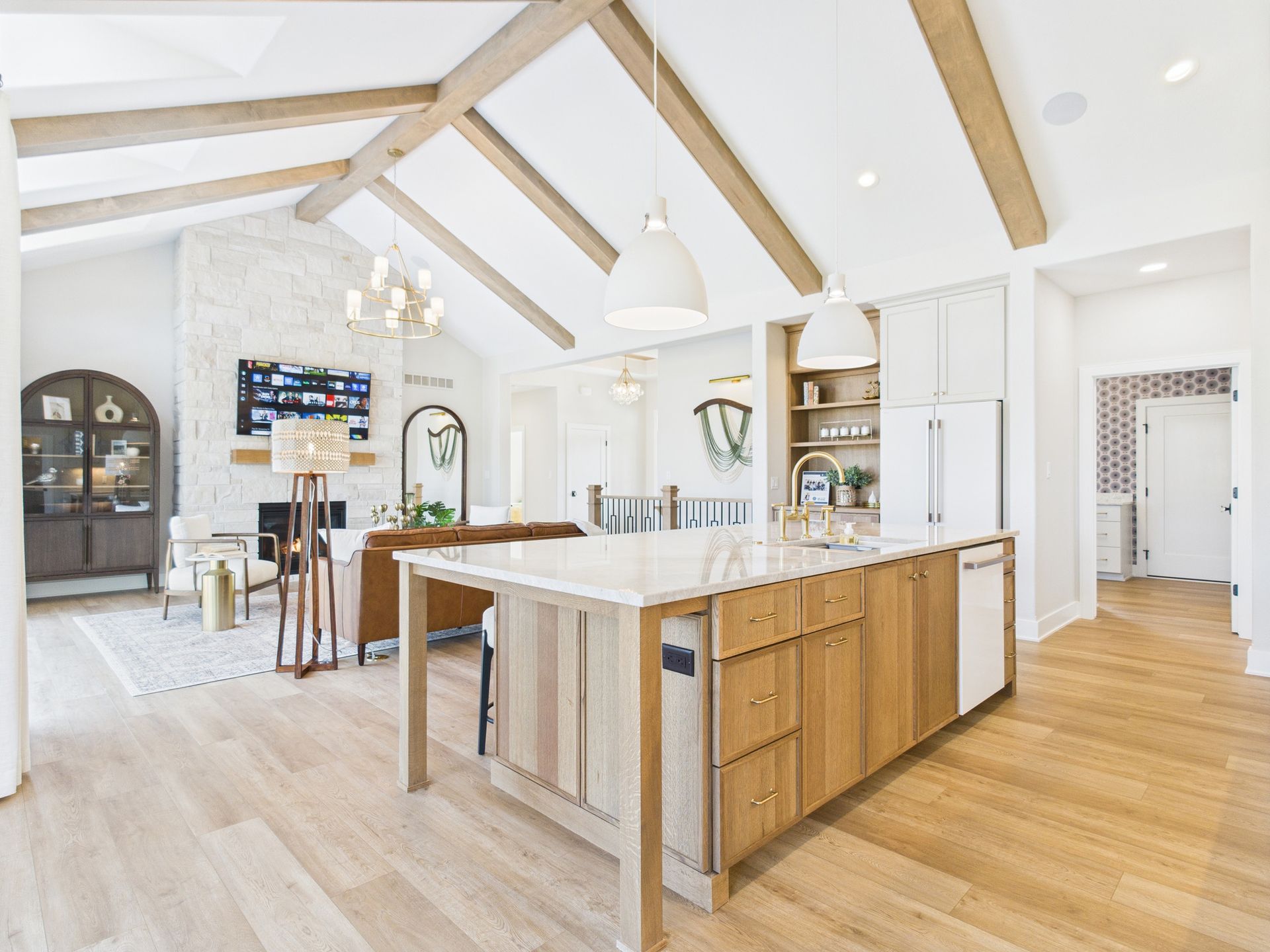 Open-concept kitchen and living area with wooden beams, island, and fireplace. White walls and light wood floors.