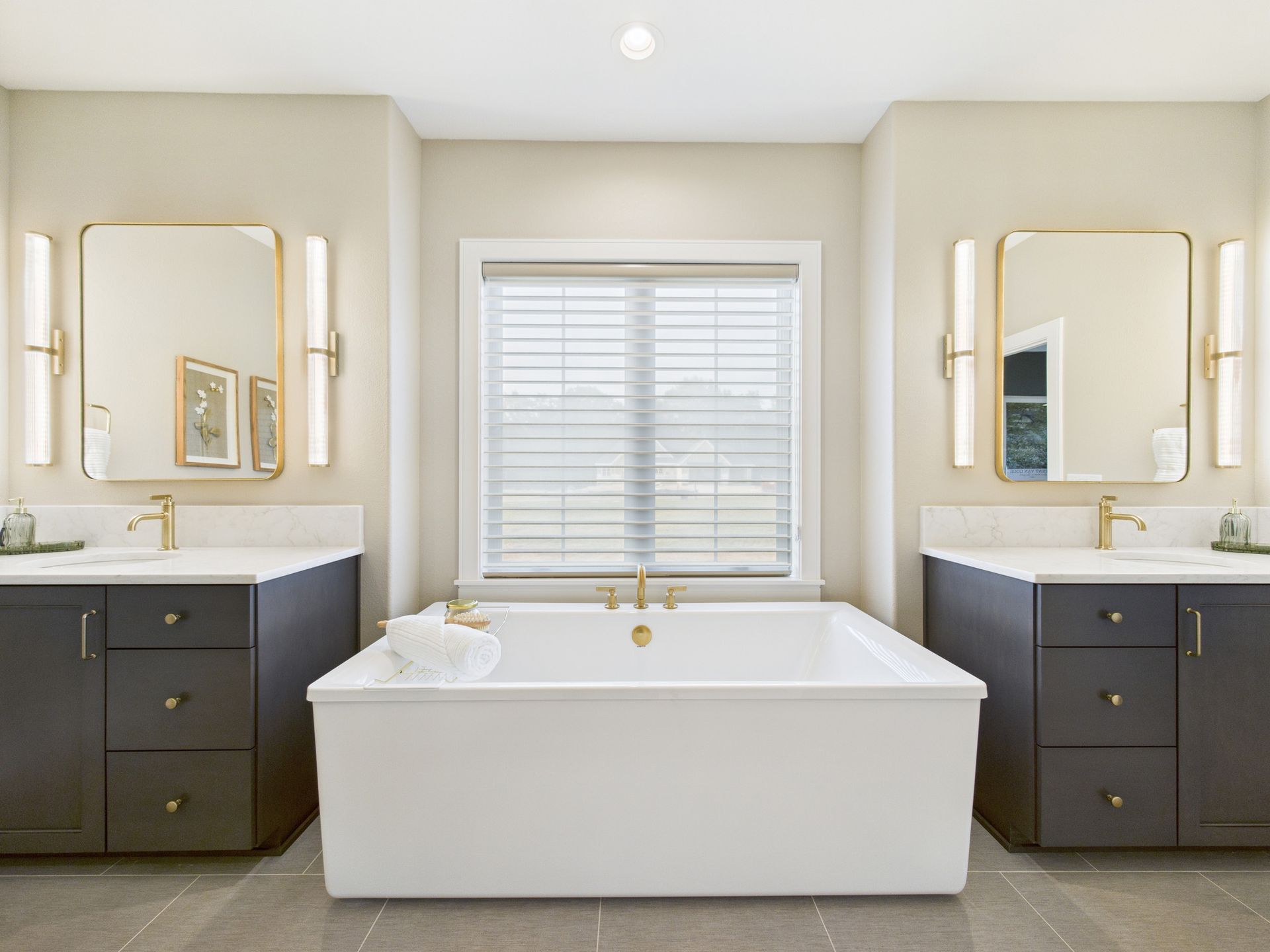 Modern bathroom with a white tub, dark gray vanities, gold fixtures, and a window.
