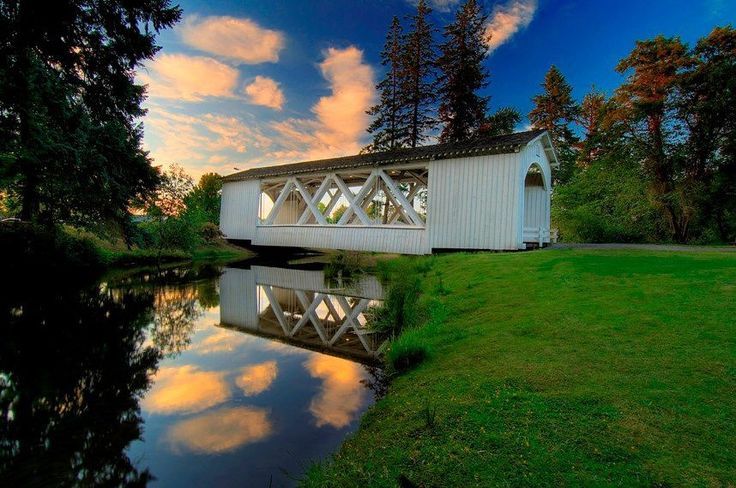 White covered bridge reflecting in calm water under a sunset sky.