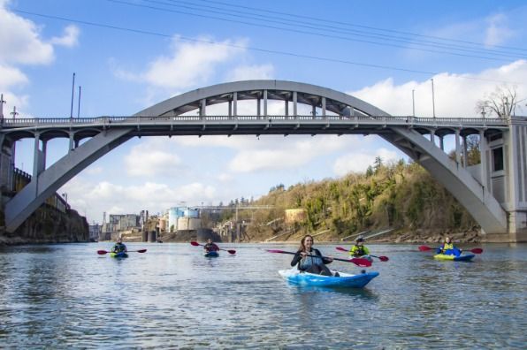 Kayakers on a river in Oregon City OR, under a large concrete bridge. Blue sky and trees in the background.