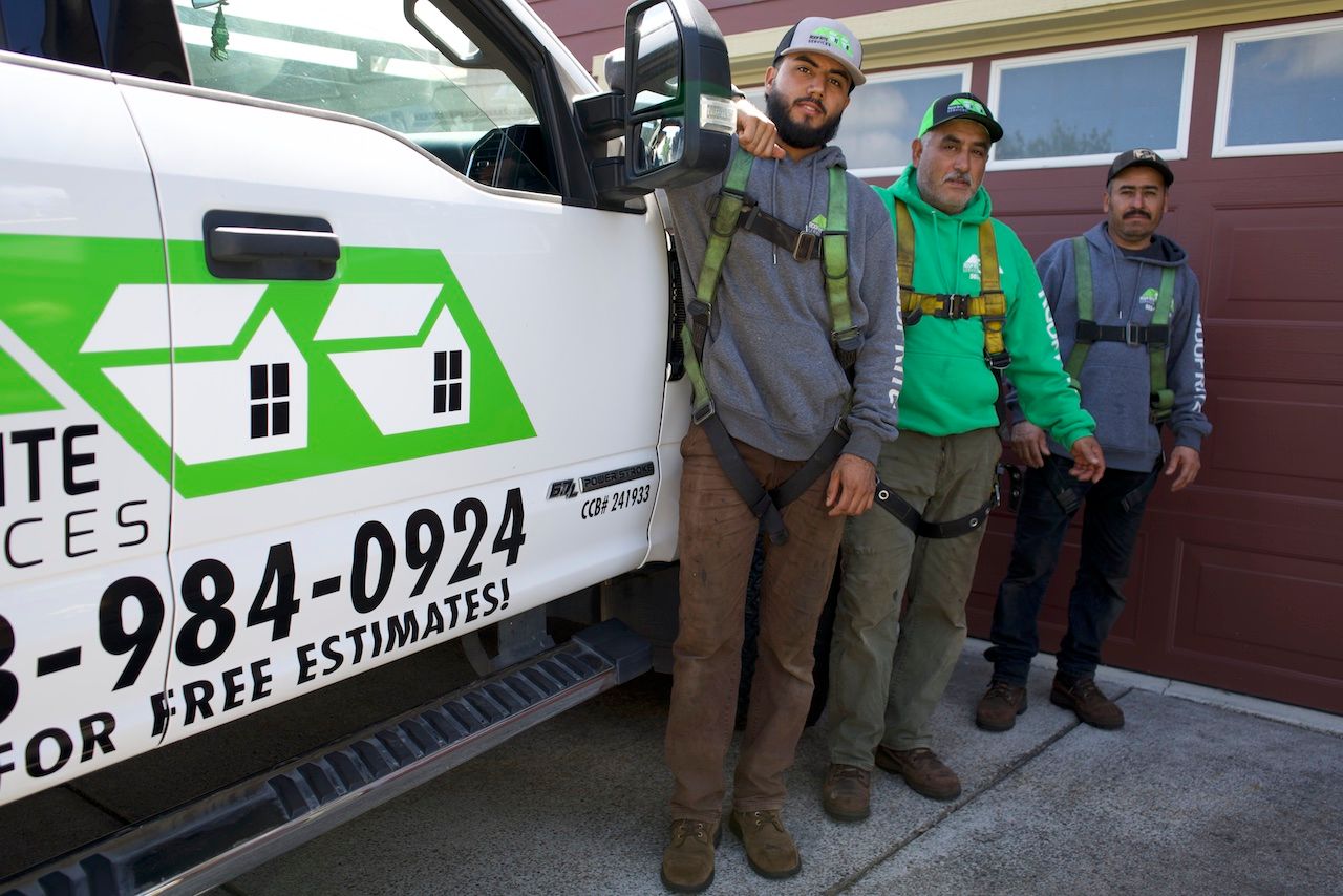 Three men in work attire stand by a truck, posing. One points, all wearing safety harnesses, smiling.