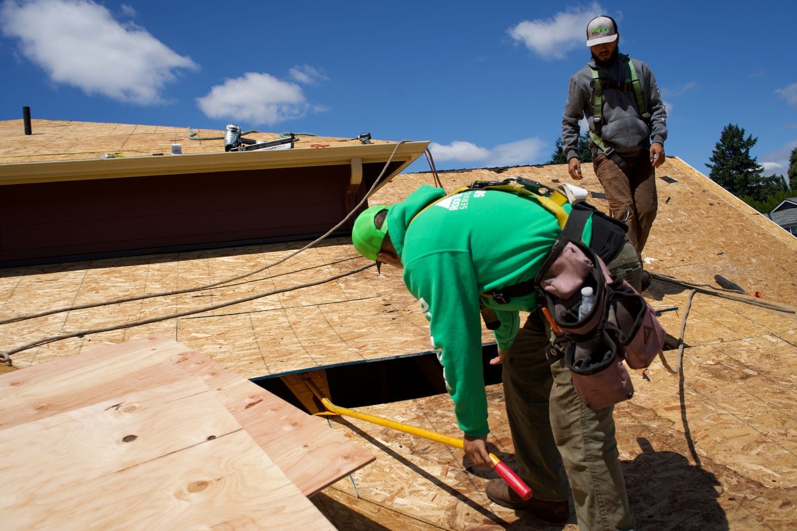 Two roofers on a roof with plywood, one looking into an opening, wearing safety gear, sunny day.