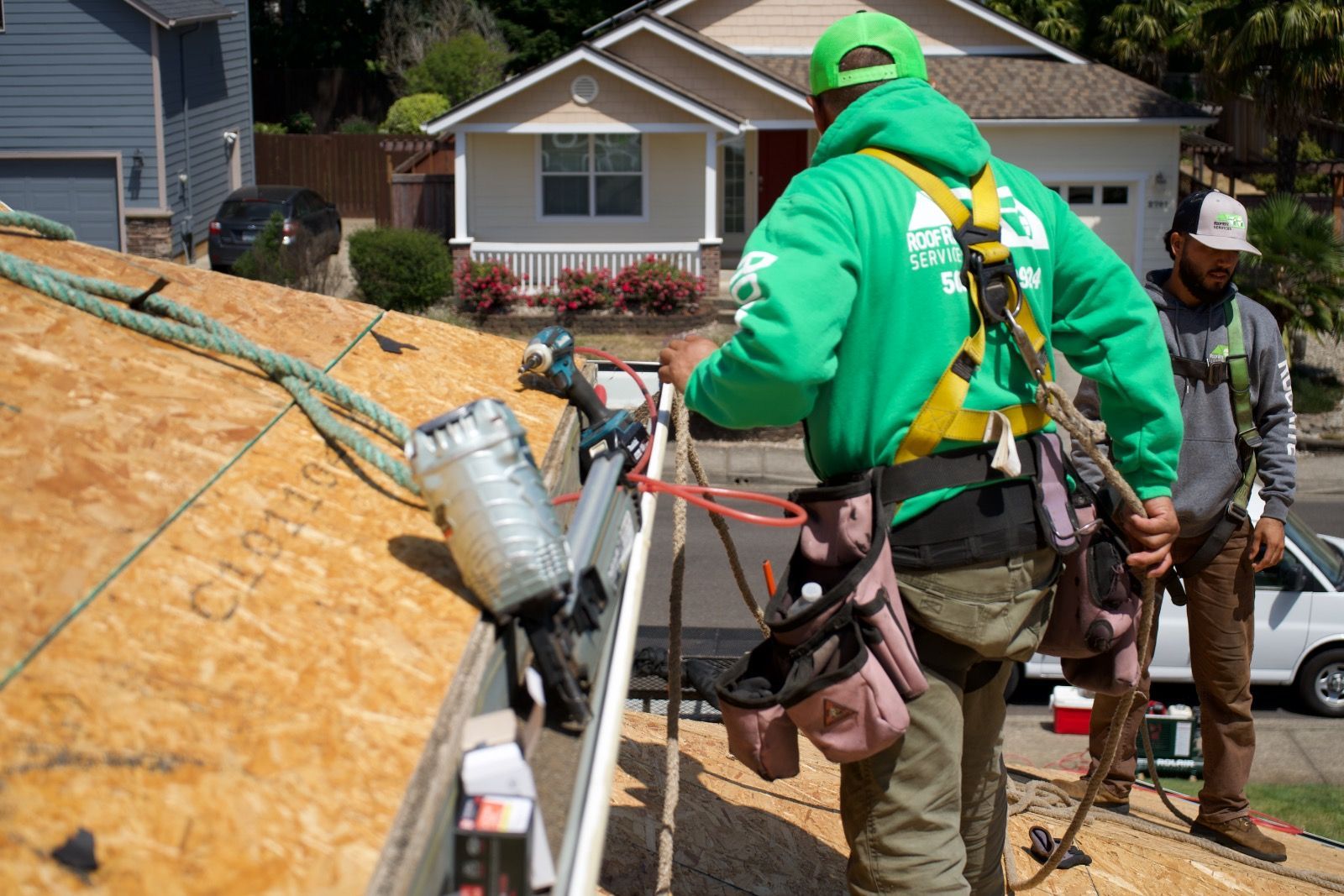 Two roofers in Salem OR, wearing safety harnesses on a rooftop, one in a green hoodie with tool belt, working on a house.