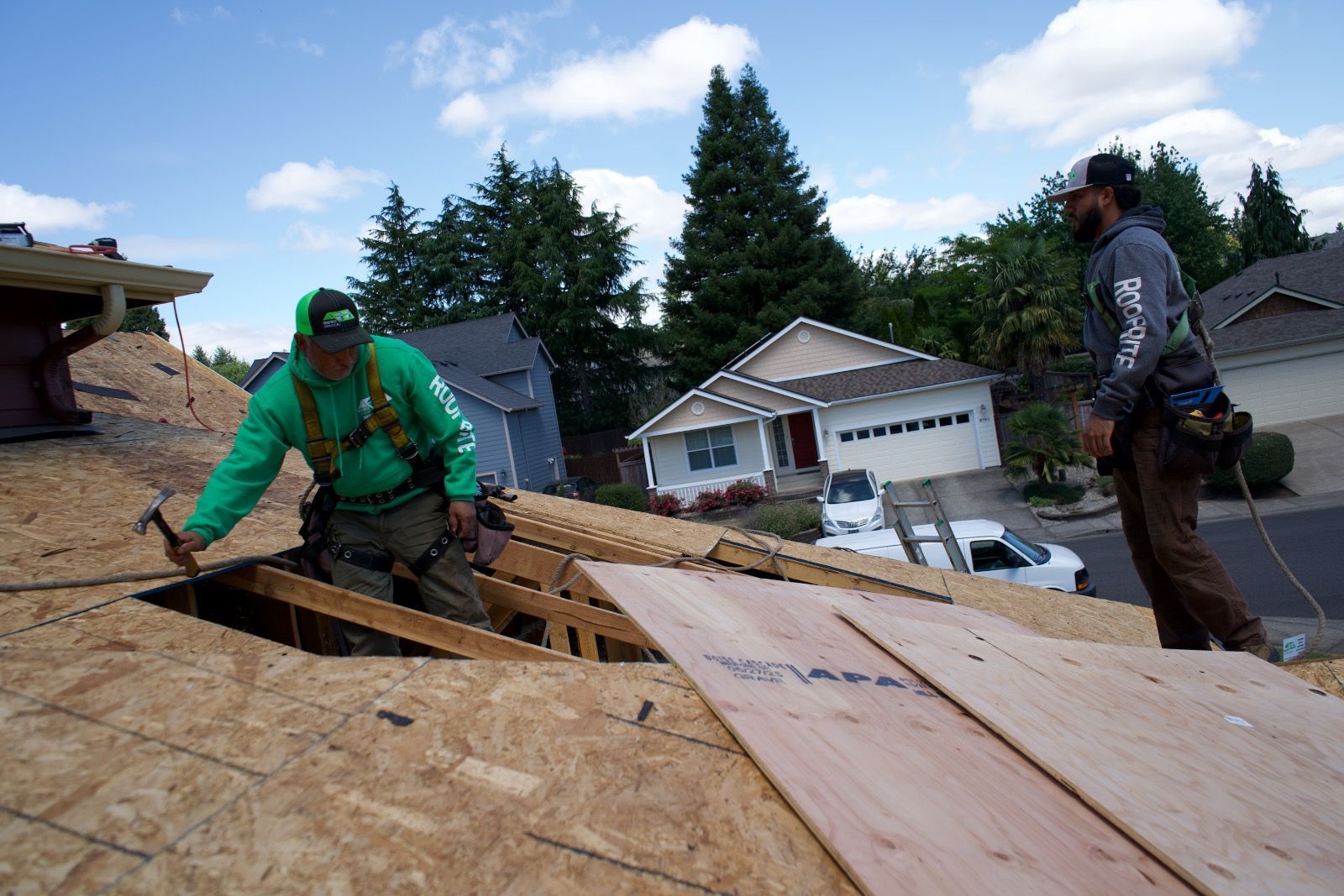 Two roofers in Salem OR working on a rooftop, with one using a hammer and the other watching. Houses and trees in background.