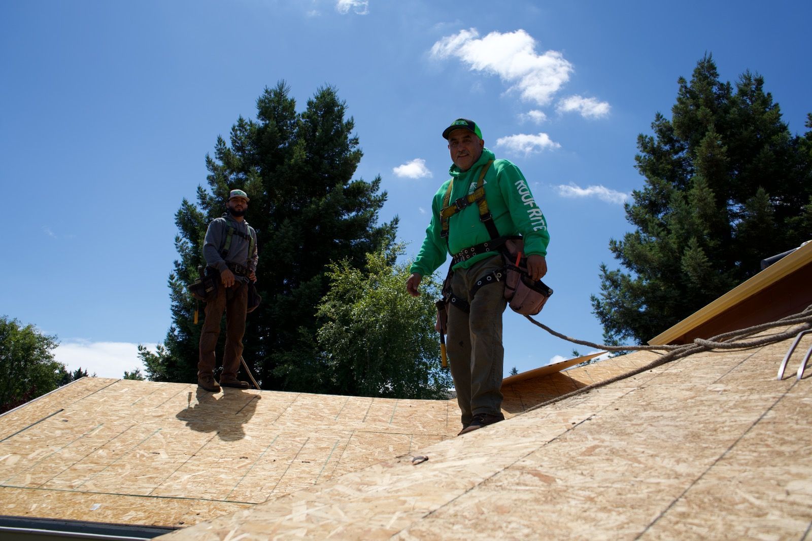 Two roofers on a partially built roof, wearing safety harnesses. Sunny day, blue sky, trees in the background.