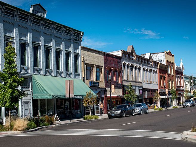Row of colorful buildings in Dallas OR along a sunny street, parked cars, clear blue sky.