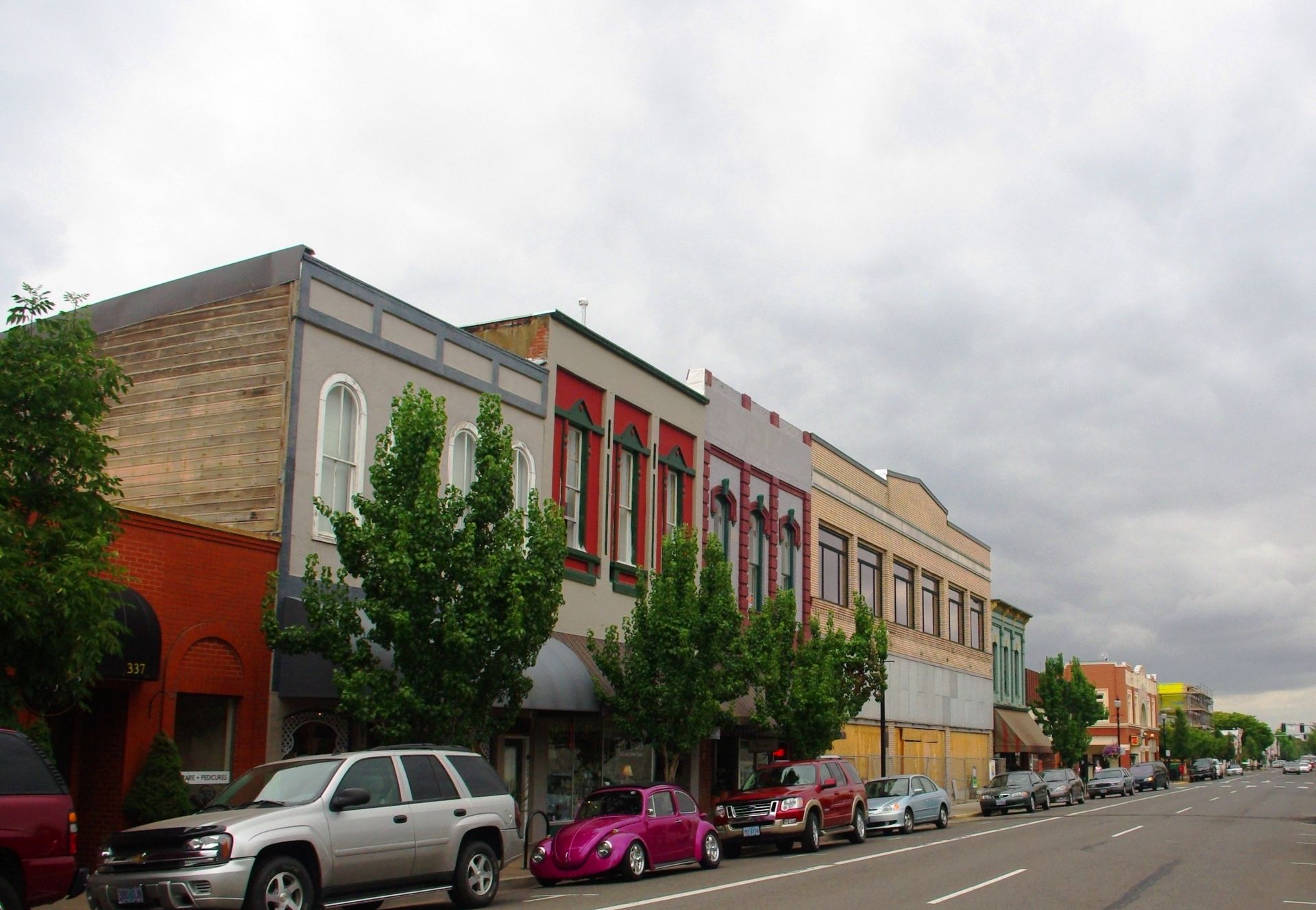 Row of colorful storefronts with cars parked in front, under a cloudy sky.