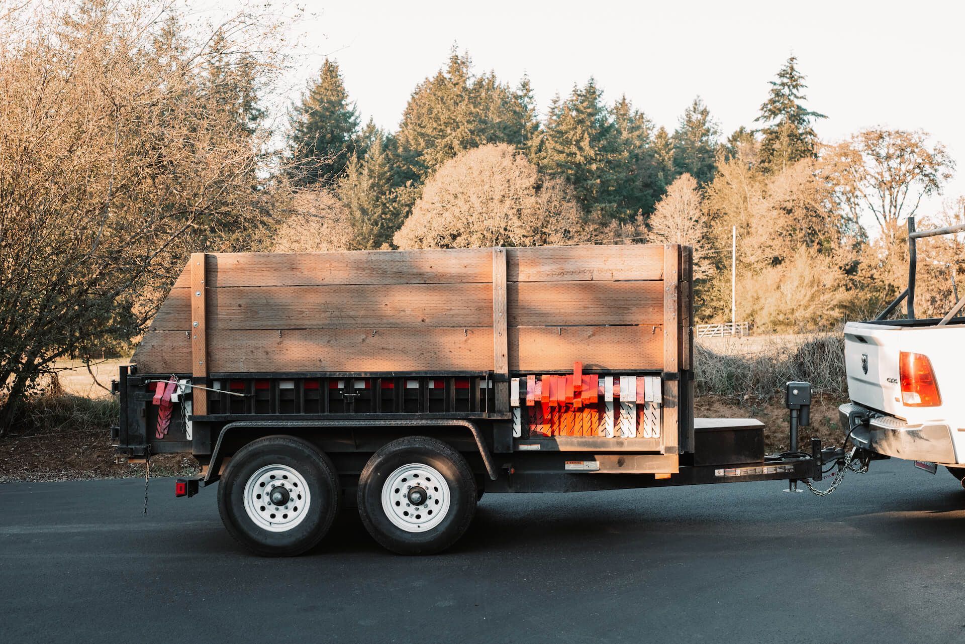Trailer with wooden walls attached to a white truck parked on asphalt with trees in the background.