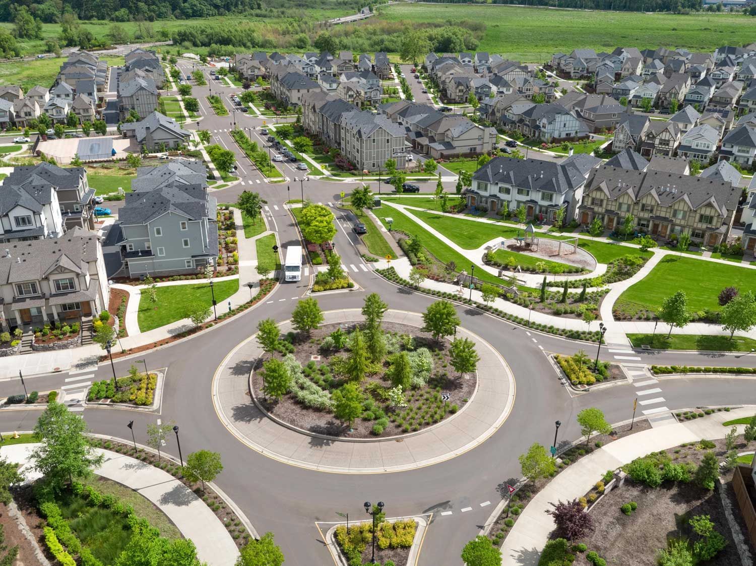 Aerial view of a suburban neighborhood with a roundabout surrounded by houses, green lawns, and trees on a sunny day.