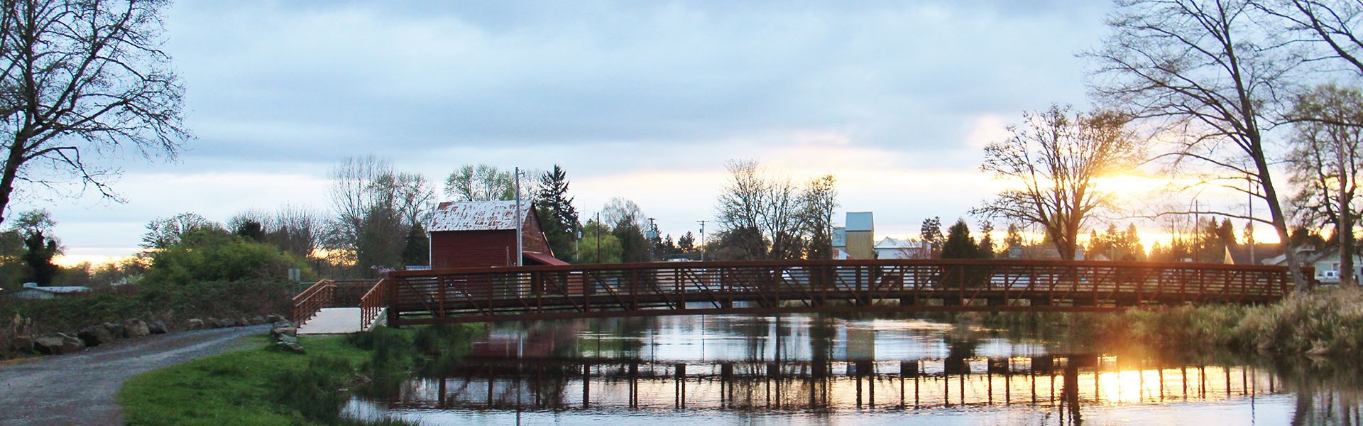 A wooden bridge spans a river at dusk, with a red building on the bank.