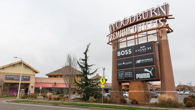 Woodburn Premium Outlets sign with brand logos, stores, and a cloudy sky.