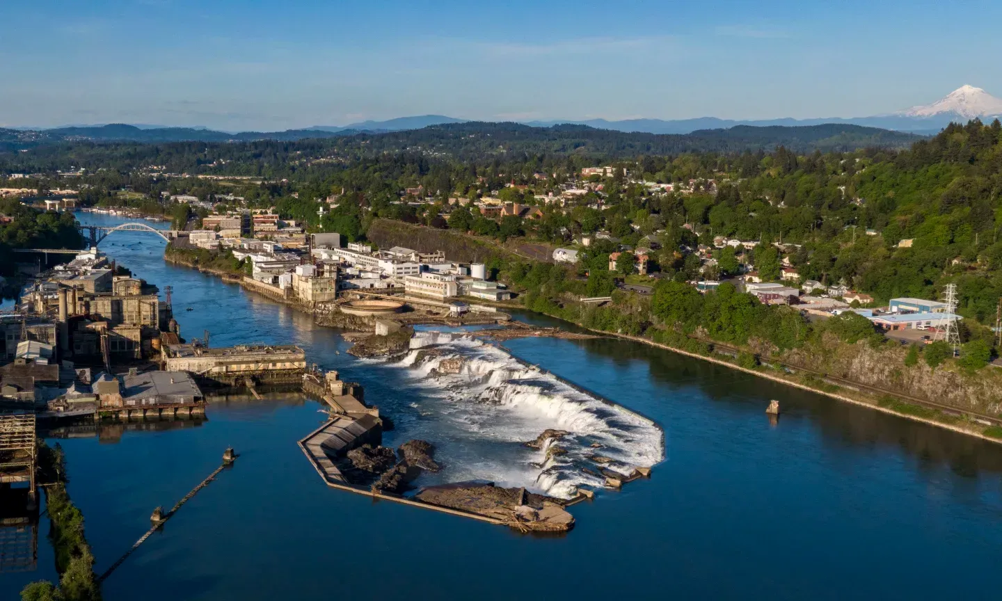 Aerial view: Willamette Falls, Oregon, with waterfall and buildings. Blue river, lush green hills, and distant mountain.