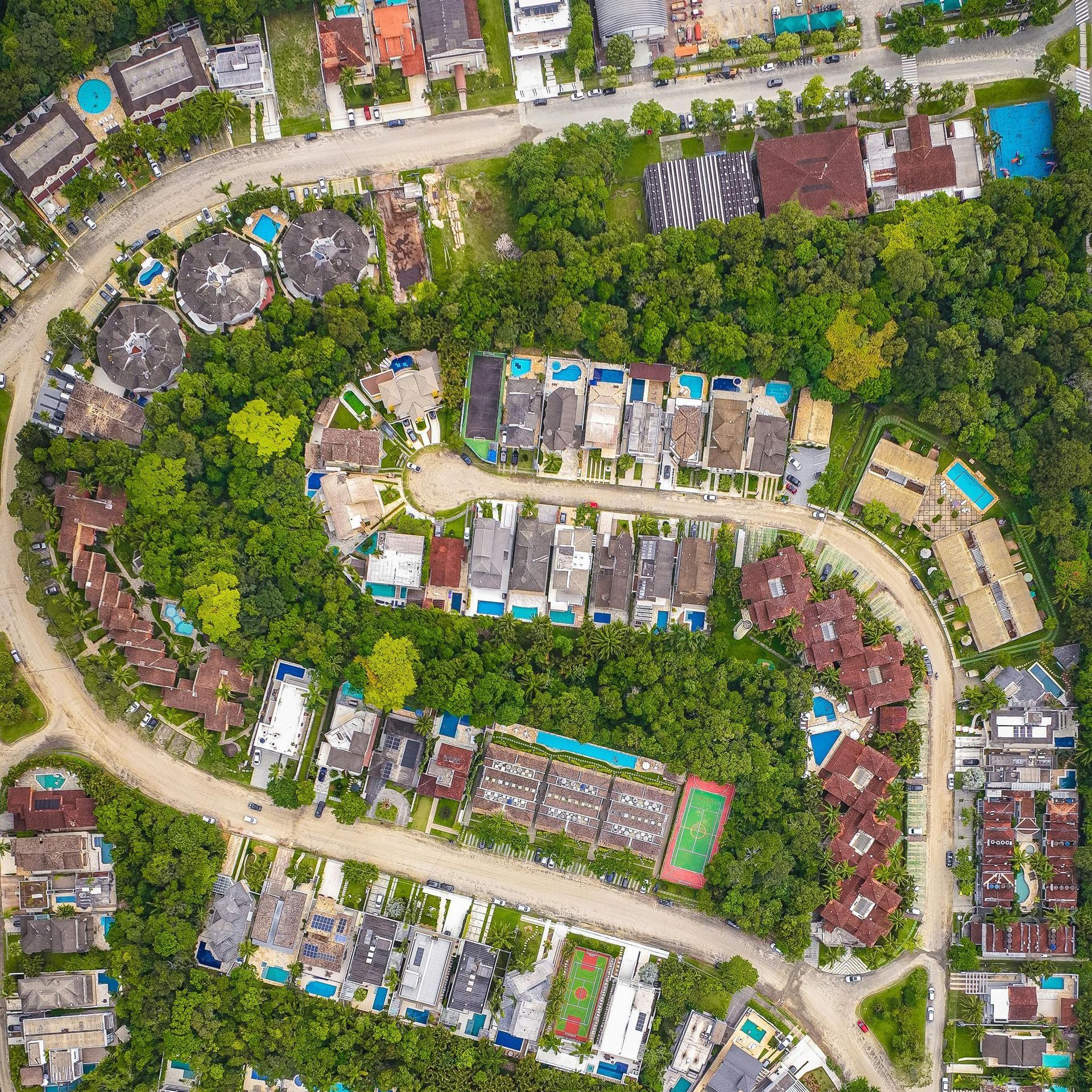 Aerial view of a winding road through a lush, green residential area with various buildings, pools, and a tennis court.