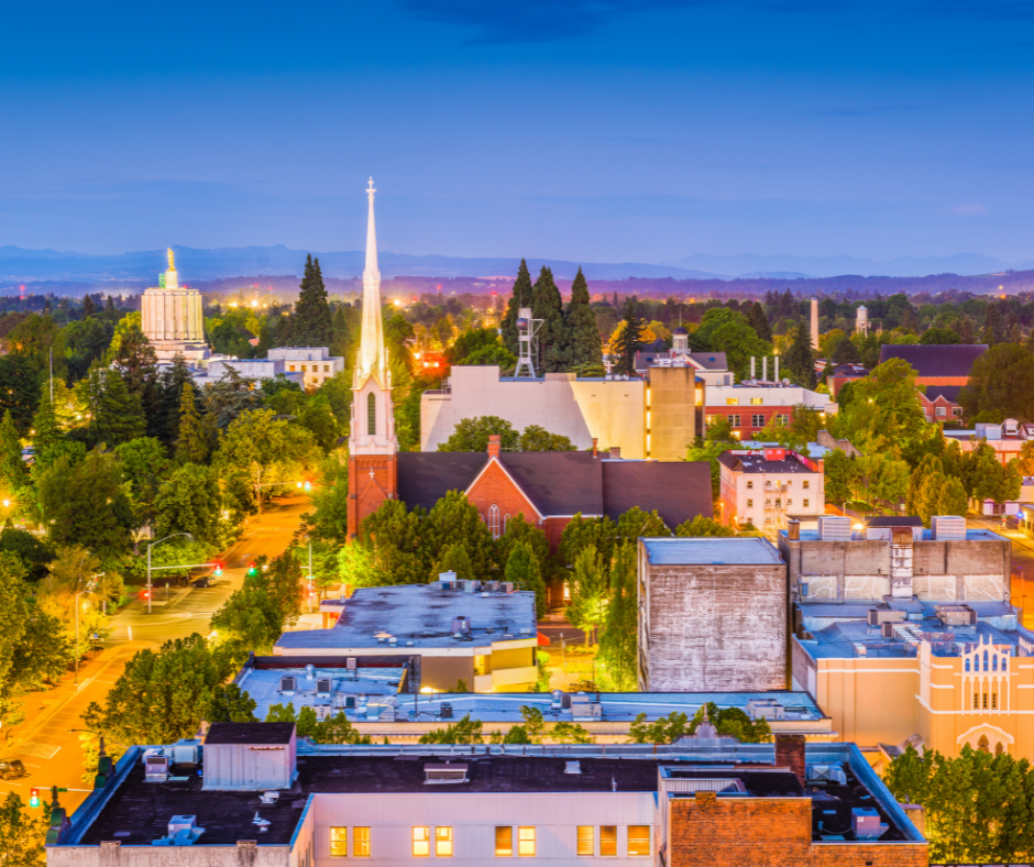City skyline at dusk with a tall church spire and surrounding buildings, trees, and a blue sky.