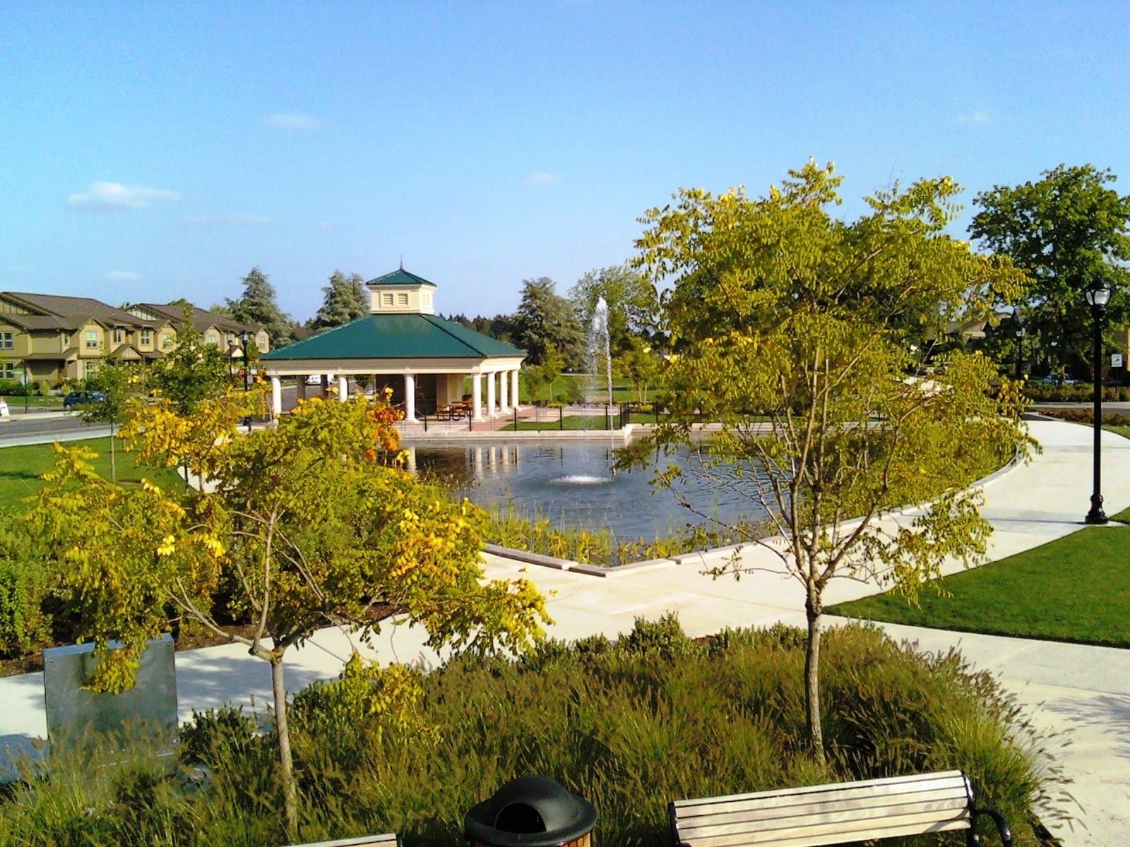 Park with gazebo, fountain, and pond under a clear blue sky. Green grass and trees surround a paved walkway.