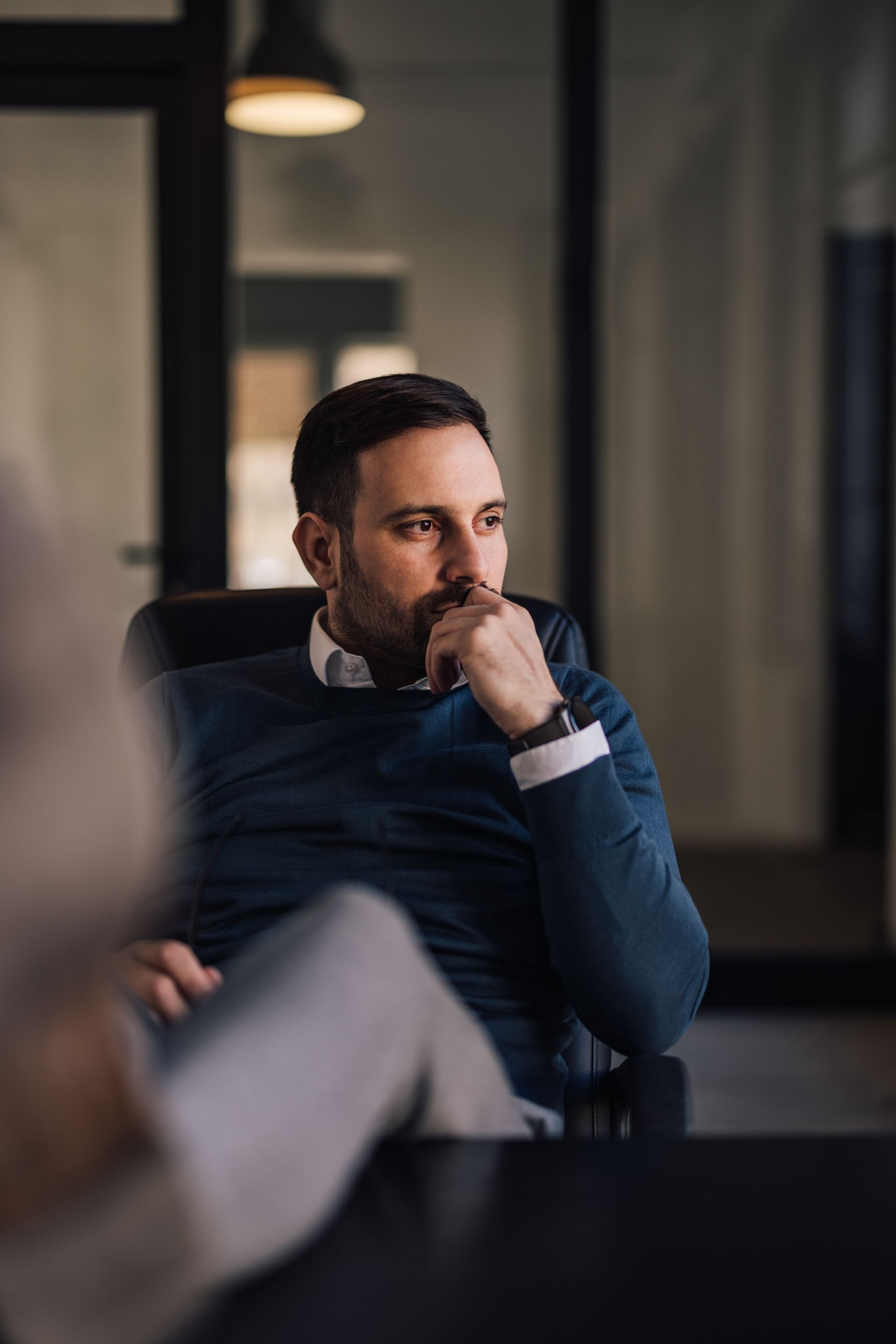 Man in a dark sweater sits thoughtfully in an office, resting his hand on his chin.