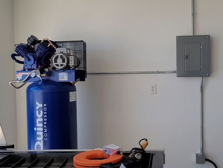 Blue air compressor in a garage beside an electrical panel, with orange hose on the floor.