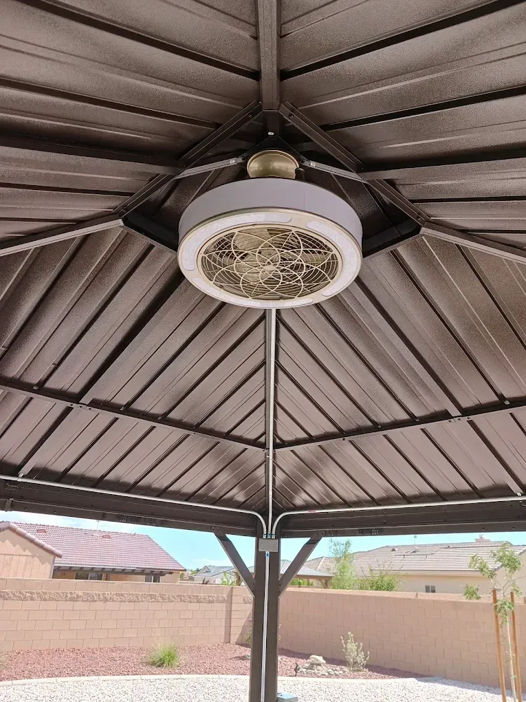 Outdoor gazebo ceiling fan with light, under a dark metal roof in a walled backyard.